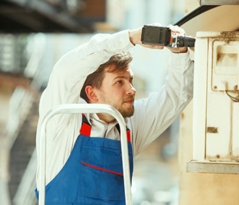 A person wearing a blue apron over a white shirt is standing on a stepladder, using a drill to install an air conditioning unit on the wall. The backdrop is slightly blurred, suggesting an outdoor setting—typical of HVAC services in Los Angeles County, CA.