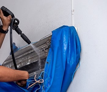 A person is meticulously cleaning an air conditioning unit mounted on a wall using a high-pressure water spray. The unit, partially covered with a blue tarp to collect water and debris, showcases the professional touch you'd expect from top HVAC services in Los Angeles County, CA.