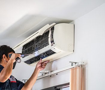 A technician in uniform expertly uses a screwdriver to repair a wall-mounted air conditioning unit. The open front panel reveals the filters and internal components, while sunlight gently streams through the curtains. Providing top-notch HVAC services in Los Angeles County, CA.