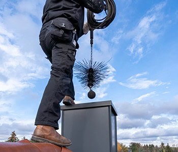 A person in black clothing and brown boots is expertly cleaning a chimney with a large brush on a roof, showcasing professional HVAC services in Los Angeles County, CA. The sky is cloudy and blue in the background.