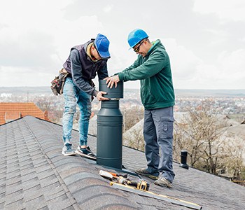 Two workers in protective gear install a vent or pipe on a residential rooftop, representing the expertise of HVAC services in Los Angeles County, CA. Surrounded by tools and wearing blue helmets, they work diligently against a backdrop of scattered trees and suburban homes.