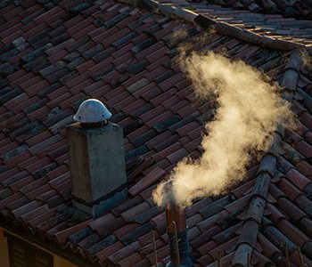 Smoke rises from a chimney on a red-tiled roof, its wispy trail curling gracefully against the backdrop of rooftops. The lighting hints at early morning or late afternoon, reminiscent of the tranquility found in Los Angeles County after expert HVAC services ensure comfort.