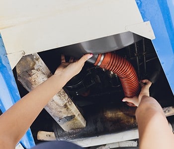 A person is diligently performing maintenance underneath a vehicle, carefully handling an orange flexible hose while working amidst the blue and metallic parts. Their skills are as essential here as in providing top-notch HVAC services in Los Angeles County, CA.