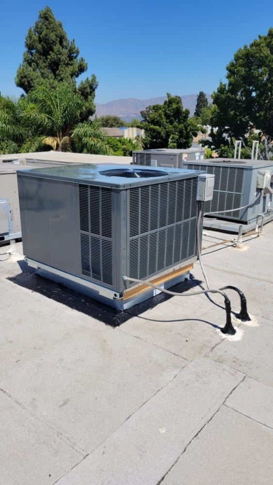 Air conditioning units installed on a flat rooftop, surrounded by trees and distant mountains under a clear blue sky.