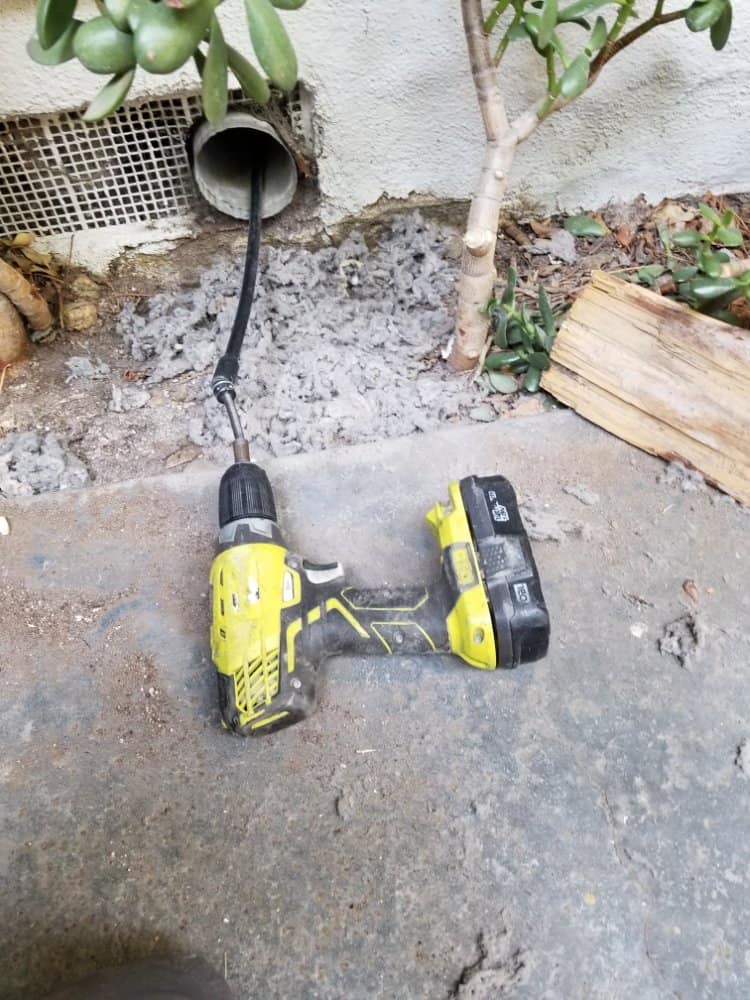 A yellow and black cordless drill lies on a concrete surface next to a plant. A cable is coming out of a nearby wall pipe covered with a mesh screen. Debris and wooden planks are visible on the ground.