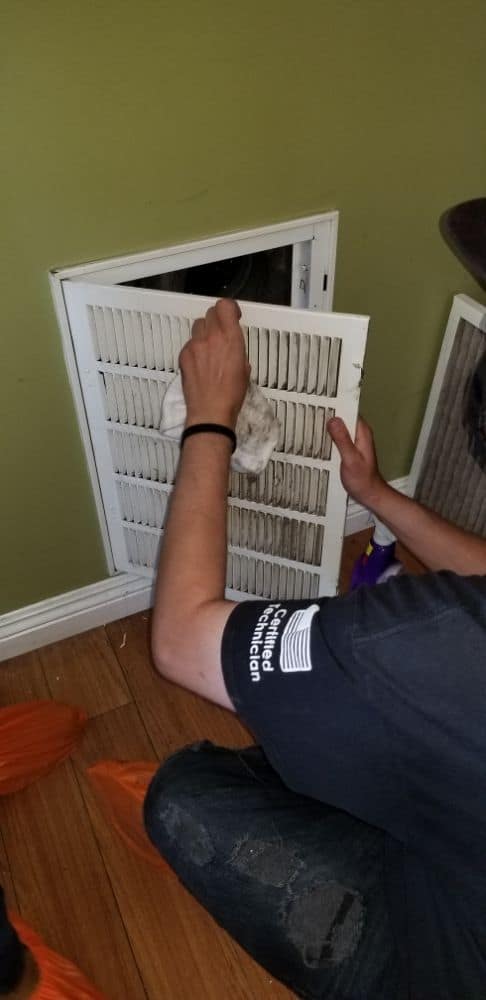 A person wearing a black shirt is cleaning a white air vent on a green wall using a cloth. The floor is wooden. The person is crouched down, focusing on the task. An orange item is partially visible on the floor.