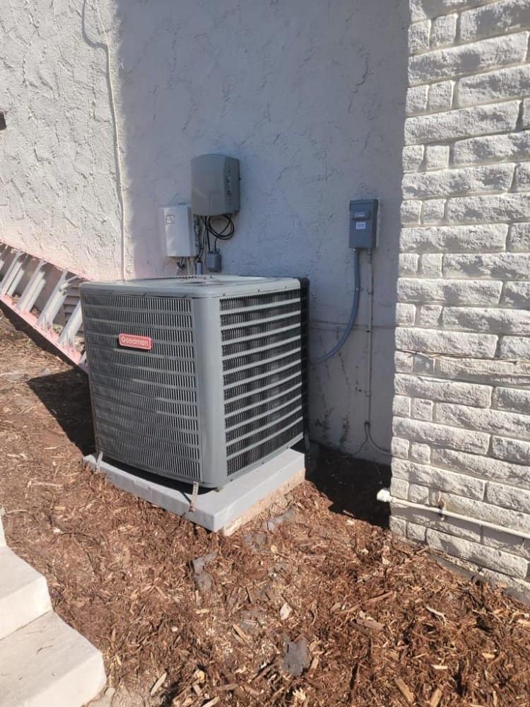 A gray air conditioning unit is installed against the exterior wall of a building. There is a white cable box mounted above it, and a hose running down the wall. A ladder is propped against the wall nearby. The ground is covered with mulch.