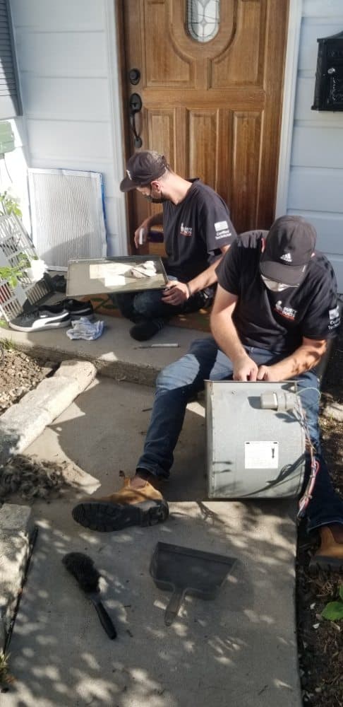 Two people in black shirts and caps are repairing equipment on a house's doorstep. One is seated, working on a metal device, while the other kneels, using tools. A doormat lies nearby, and a wooden door is in the background. Various tools are scattered around.