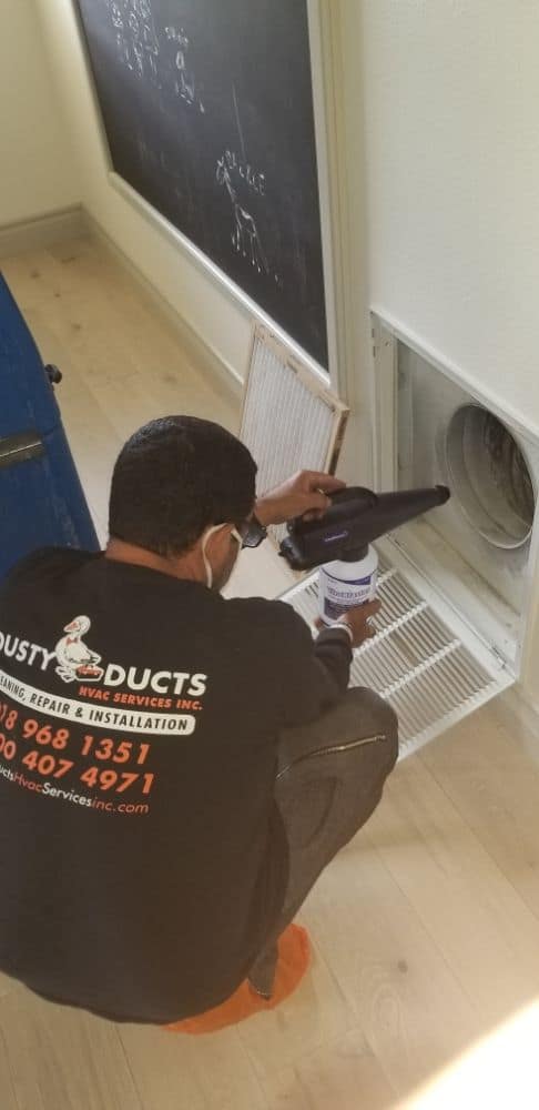 A technician in a black shirt and face mask is cleaning an air duct with a vacuum tool. He is kneeling on a light wood floor next to an open vent cover. A chalkboard is partially visible on the wall behind him.