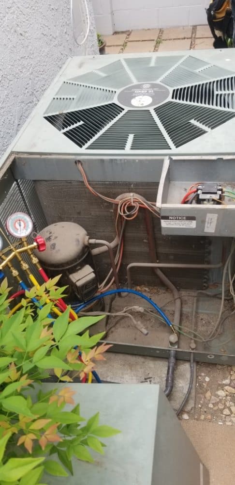 An open air conditioning unit with visible wires, pipes, and components. A nearby shrub with green leaves is partially in view. The unit is situated against a light-colored wall on a patio with paving stones.