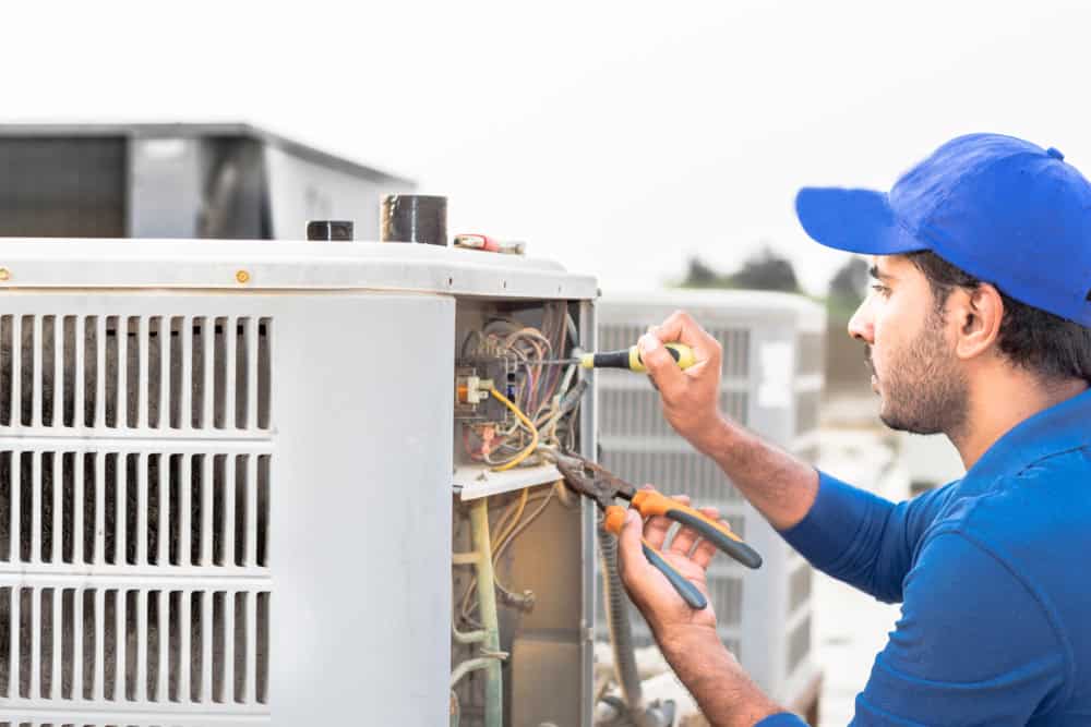 A technician in a blue cap and shirt expertly uses a screwdriver to repair an outdoor air conditioning unit. Providing top-notch HVAC services in Los Angeles County, CA, he holds pliers in his other hand while diligently working on exposed wires of the unit.