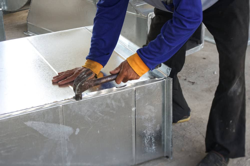 A skilled individual in a blue and yellow long-sleeve shirt expertly wields a hammer on a metal sheet, capturing the essence of hands-on craftsmanship in an HVAC services Los Angeles County CA workshop, with metal pieces scattered artfully in the background.