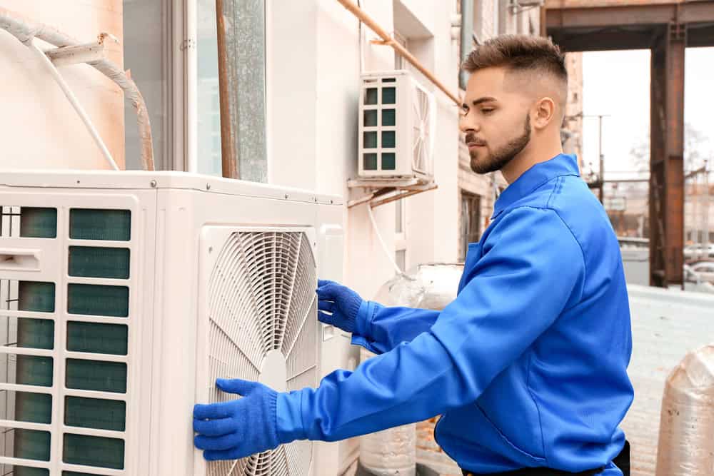 A technician in a bright blue uniform and gloves is expertly installing an outdoor air conditioning unit, showcasing the precision typical of HVAC services in Los Angeles County, CA. He is focused on the task against a vibrant urban backdrop of buildings and industrial pipes.