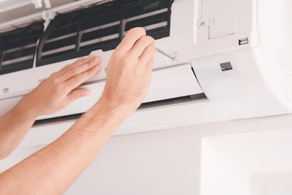 A person is using a screwdriver to repair or install a wall-mounted air conditioner in Los Angeles County, CA. One hand holds the tool, while the other steadies the unit. The air conditioner's cover is open, revealing its interior components—a touch of expert HVAC services at work.