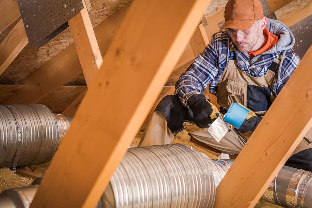 A worker in overalls and a cap meticulously applies insulation tape to metal ductwork in an attic with visible wooden beams and a sloped ceiling. With safety goggles on, he represents the skilled professionals of HVAC services Los Angeles County, CA, using a brush for precision in his craft.