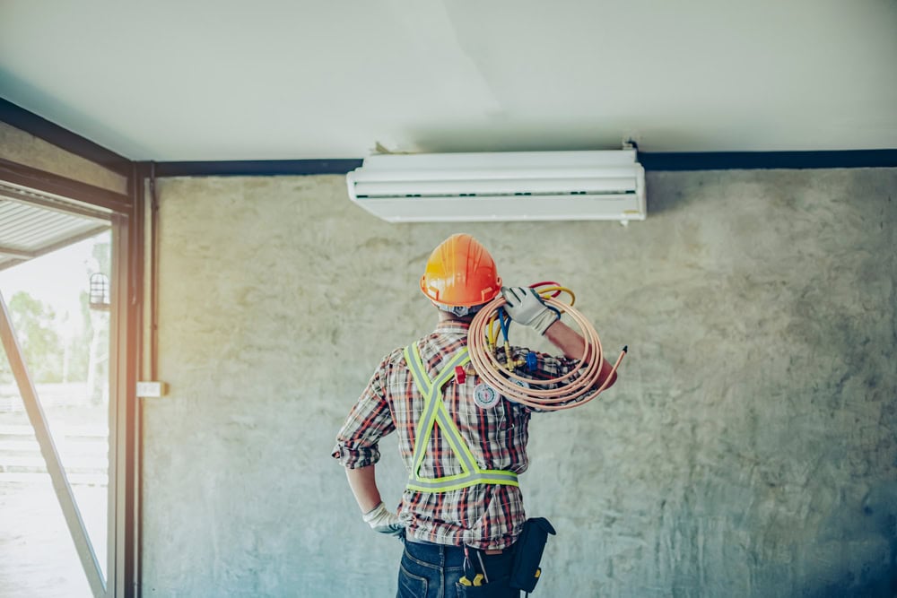 A construction worker in a plaid shirt and orange hard hat is inspecting an air conditioning unit on a wall, delivering expert HVAC services in Los Angeles County, CA. They hold a coil of wires, wearing a tool belt and safety harness in the room with bare concrete walls.