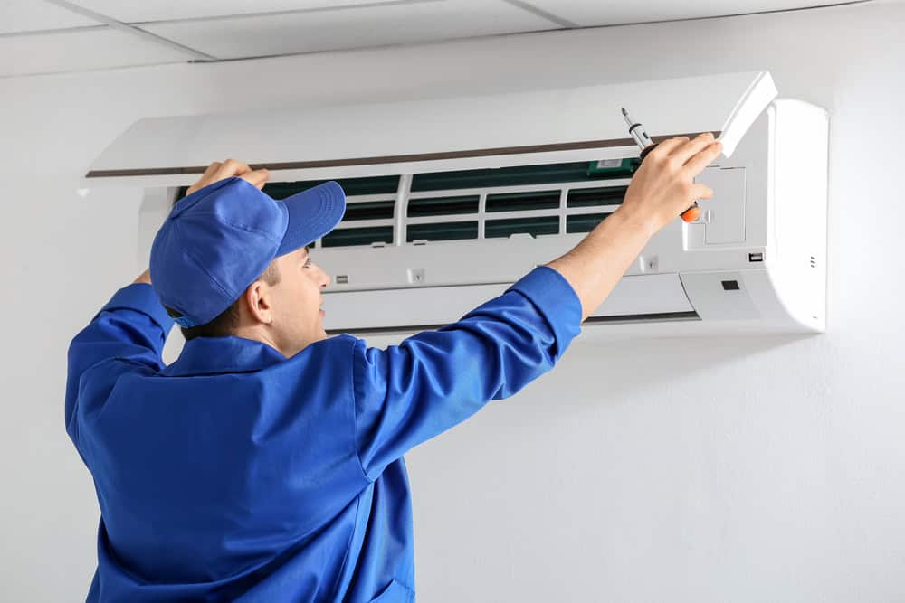 A technician in a blue uniform and cap expertly provides HVAC services, skillfully repairing or installing a wall-mounted air conditioning unit. In an indoor setting, he uses a screwdriver to work on the open panel, showcasing his expertise right here in Los Angeles County, CA.