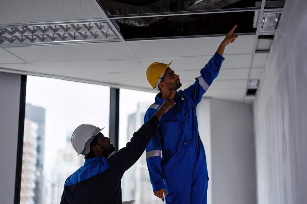 Two construction workers wearing hard hats and blue overalls inspect a ceiling in a bright office space, ensuring quality HVAC services. One points upward while the other observes. The office has large windows overlooking a Los Angeles County cityscape.