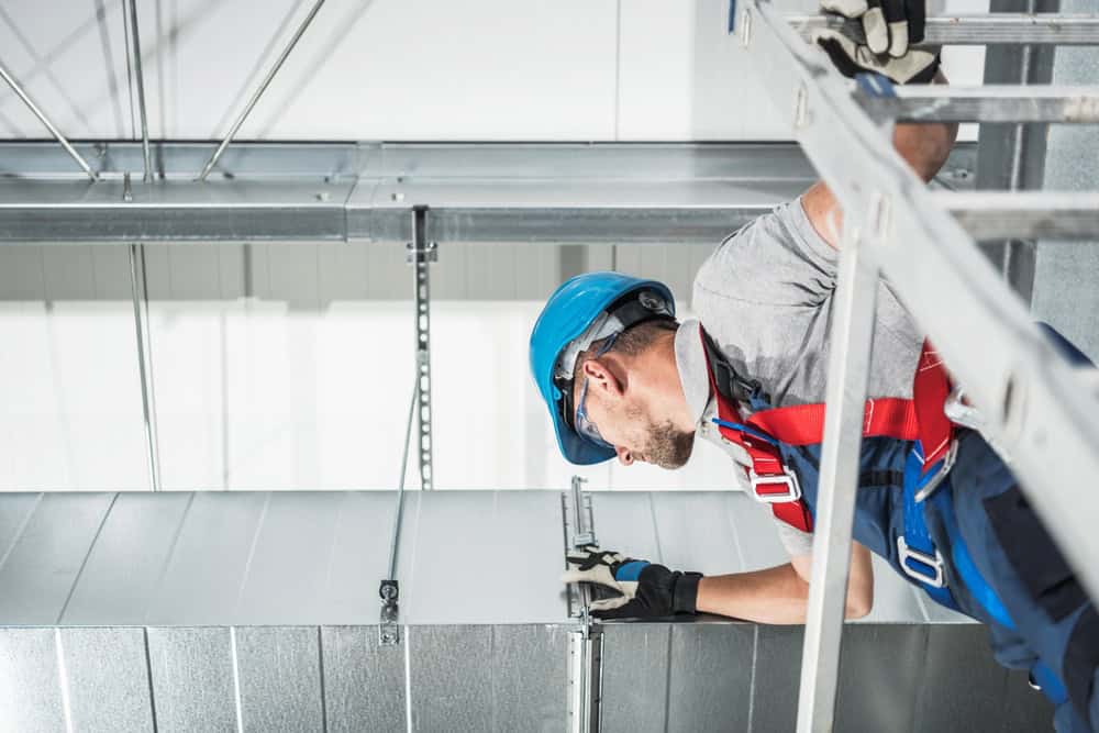 A worker in a blue hard hat and safety gear is perched on a ladder, inspecting ductwork in an industrial setting. Surrounded by metal ducts and beams, he delivers exceptional HVAC services in Los Angeles County, CA, with unwavering focus and dedication to the task.